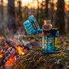 A translucent blue Blazer CG-001 Cigar Torch Lighter with a black and silver lighter mechanism, shown open on a mossy rock outdoors with a campfire and forest in the background.