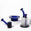 A set of blue and clear glass smoking accessories arranged on white display blocks against a white studio background. The set includes a blue glass mouthpiece, a black textured glass bowl, a blue ring holder, and a larger clear cut-glass glass with a blue neck and stopper on a round base.