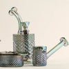 A set of iridescent glass smoking accessories, including a bowl, a small pipe, a larger bowl with a diamond-cut textured pattern, and a glass mouthpiece, displayed on a clean off-white background.