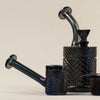 A set of dark iridescent glass smoking accessories, including a bowl, a pipe, a small cup, and a large decanter, all featuring a textured diamond-cut pattern and a glossy finish, set against a plain beige background.