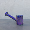 A small metallic smoking pipe with an iridescent purple and blue finish, featuring a textured knurled bowl and a straight stem, resting on a gray surface against a textured gray background.
