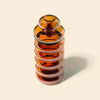 An amber glass bong with a matching screw-top lid, featuring a stacked design of nested ribbed glass rings, set against a solid cream background with a soft shadow.