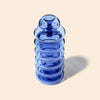 A translucent cobalt blue glass bong with a ribbed cylindrical body and a rounded stopper, shown on a warm beige background with a soft shadow.