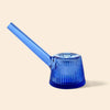 A NWTN Home translucent cobalt blue glass pipe with a ribbed, flared base and a long angled stem, shown on a solid light beige background.