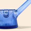 A NWTN Home close-up of a translucent cobalt blue glass water pipe with a ribbed base and a diagonal glass mouthpiece, set against a solid light beige background.