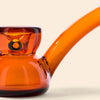 Close-up of an amber orange glass water pipe by NWTN Home with a curved stem and bowl, shown against a light cream background.