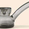 A close-up of a translucent smoky gray glass water pipe by NWTN Home with a curved mouthpiece and bowl, set against a solid light beige background.