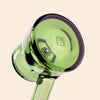 A close-up of a translucent green glass water pipe by NWTN Home with a circular bowl and stem, featuring a small 'h' logo on the bowl, set against a solid pale beige background.