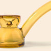 A close-up of a translucent amber glass water pipe by NWTN Home with a curved stem and bowl, shown against a plain off-white background.