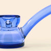 A close-up of a translucent cobalt blue glass water pipe by NWTN Home with a curved stem, set against a solid light beige background.