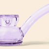 A close-up of a translucent lavender purple glass water pipe by NWTN Home with a curved bowl and long curved neck, set against a solid pale beige background.