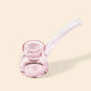 A small pink glass water pipe by NWTN Home with a rounded bowl and a curved stem, photographed on a plain warm beige background.
